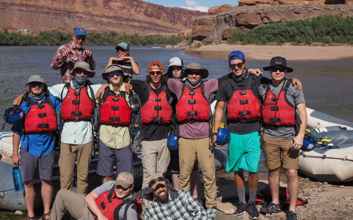 A group of students wearing life jackets wrap their arms around each other and smile for a photo on the shore of a river. There are tall canyon walls in the background.